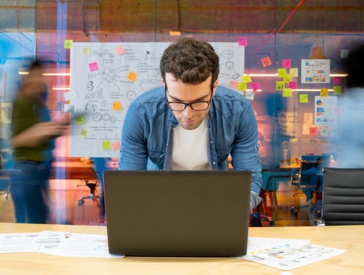 Man working at a creative office using his computer and people moving at the background